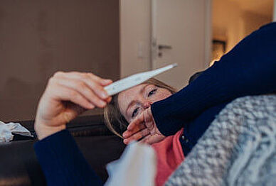Frau liegt mit Fieberthermometer in der Hand, in Decke eingehüllt auf dem Sofa