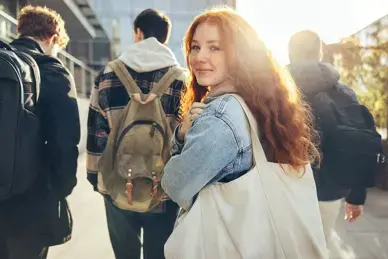 Ein jugendliches Mädchen mit langen roten Haaren dreht sich im Gehen zum Betrachter um, vor ihr gehen drei gleichaltrige Jungen