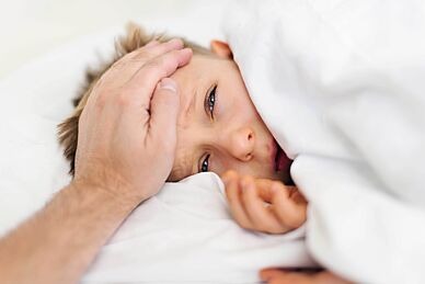 Kranker Junge liegt unter der Bettdecke, eine erwachsene, männliche Hand fühlt auf seiner Stirn die Temperatur