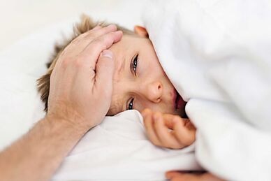 Kranker Junge liegt unter der Bettdecke, eine erwachsene, männliche Hand fühlt auf seiner Stirn die Temperatur