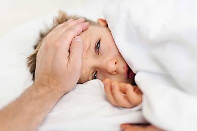 Kranker Junge liegt unter der Bettdecke, eine erwachsene, männliche Hand fühlt auf seiner Stirn die Temperatur