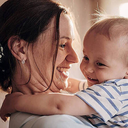 Frau hält ein Baby im Arm, beide lachen sich an