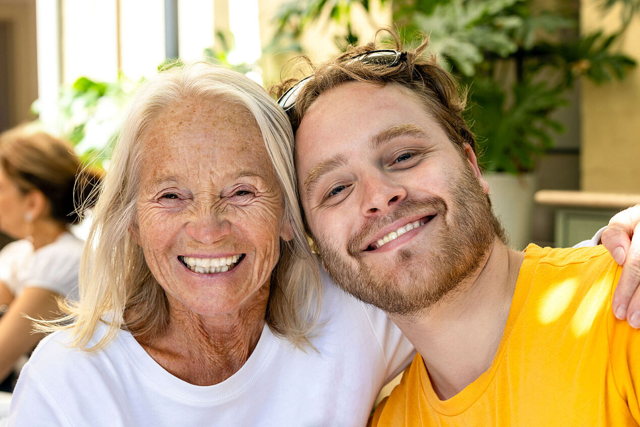 Ältere Frau legt ihren Arm um einen jüngeren Mann, beide lächeln in die Kamera