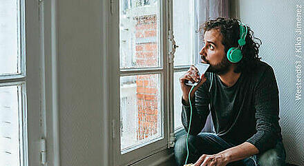 Junger Mann mit Kopfhörern und Smartphone schaut aus dem Fenster und isst nebenbei