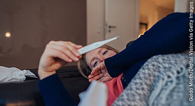 Frau liegt mit Fieberthermometer in der Hand, in Decke eingehüllt auf dem Sofa