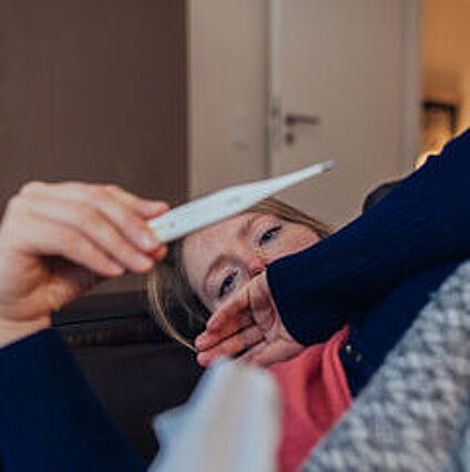 Frau liegt mit Fieberthermometer in der Hand, in Decke eingehüllt auf dem Sofa