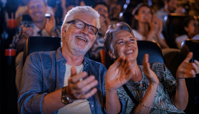 Ein Mann und eine Frau fortgeschrittenen Alters sitzen im Theaterpublikum, lachen und klatschen