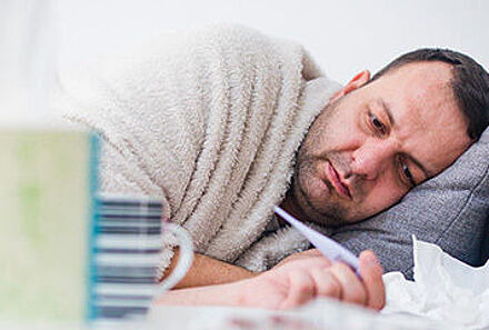 Mann liegt im Bett, hält ein Fieberthermometer in der Hand