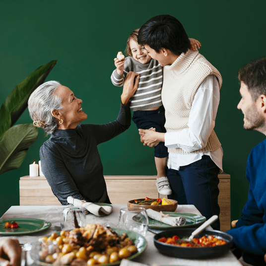 Drei Personen sitzen um einen gedeckten Tisch, eine stehende Person hat ein Kleinkind auf dem Arm. Alle schauen zum Kind.