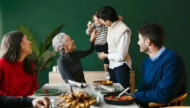 Drei Personen sitzen um einen gedeckten Tisch, eine stehende Person hat ein Kleinkind auf dem Arm. Alle schauen zum Kind.