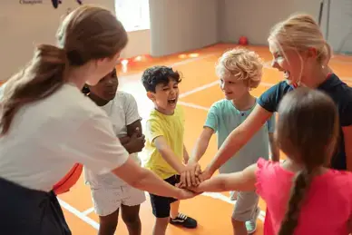 In einer Turnhalle stehen mehrere Kinder und zwei Trainerinnen im Kreis, legen in der Mitte ihre Hände aufeinander und rufen etwas