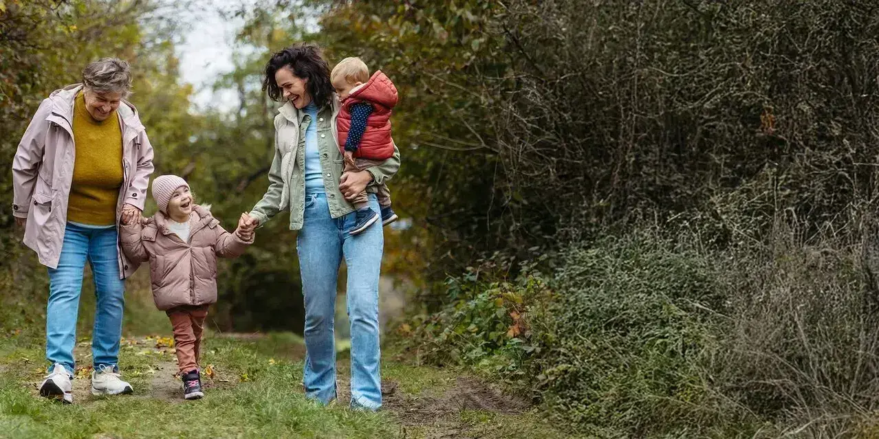 Eine ältere Dame, eine mittelalte Frau mit einem Kleinkind auf dem Arm und ein Kind gehen im Wald Hand in Hand spazieren