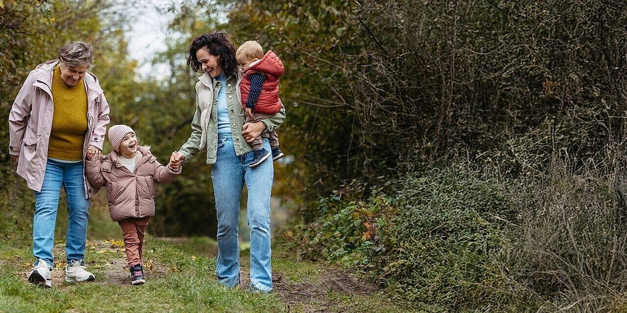 Eine ältere Dame, eine mittelalte Frau mit einem Kleinkind auf dem Arm und ein Kind gehen im Wald Hand in Hand spazieren