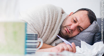 Mann liegt im Bett, hält ein Fieberthermometer in der Hand